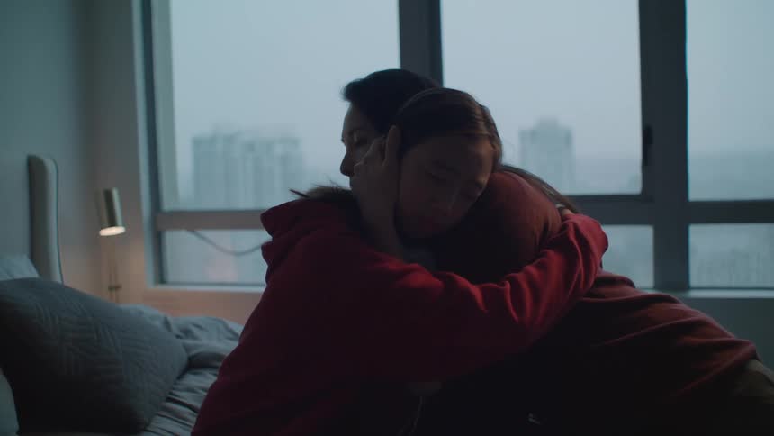 Girl sitting on a bed wearing headphones is consoled by her mother