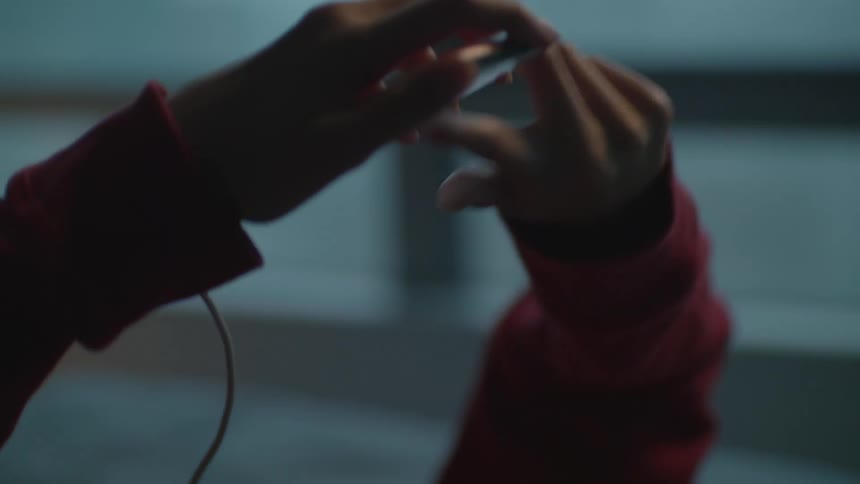 A girl in a room with headphones listening to a smart phone with a wall and window in the background