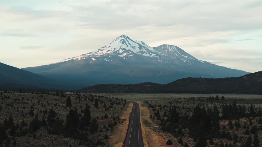 An open road with a snow covered mountain in the distance under a cloudy sky