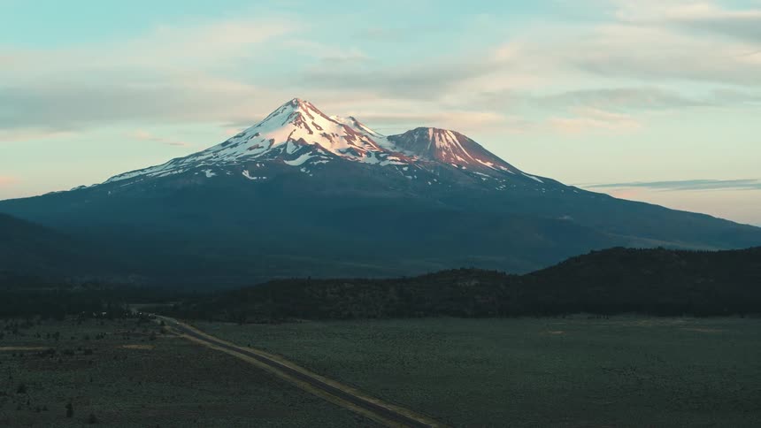 Vehicle drives along road through fields with snow capped mountain on horizon covered in mist as sunlight just touching the summit