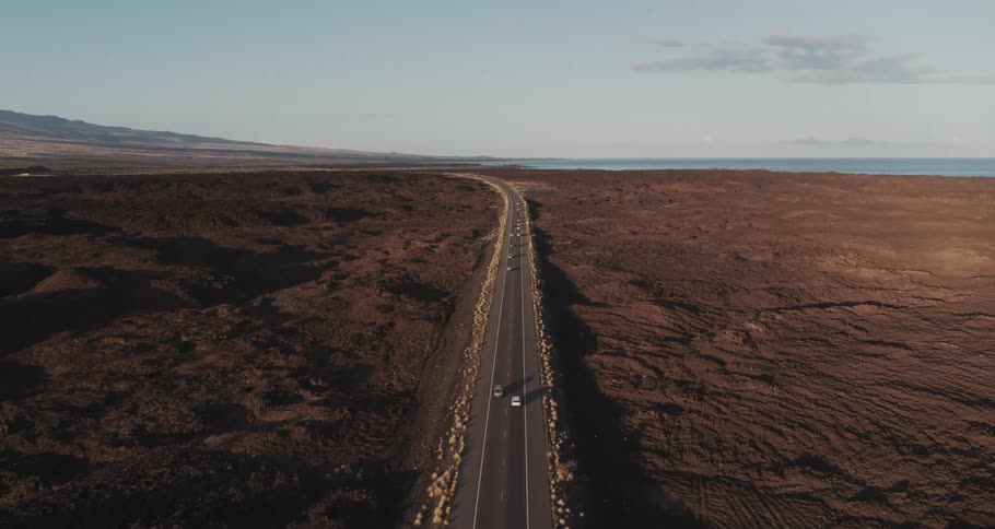 Cars and trucks travel on a long stretch of road through barren land towards the shore