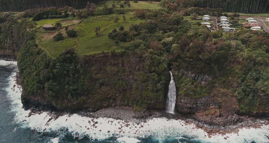 A cliff near a beach with a waterfall with trees and houses on top of the cliff