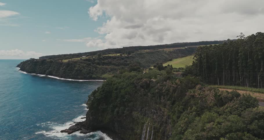 Work truck drives along highway bordering forest and cliff side waterfall flowing onto shore as waves crash