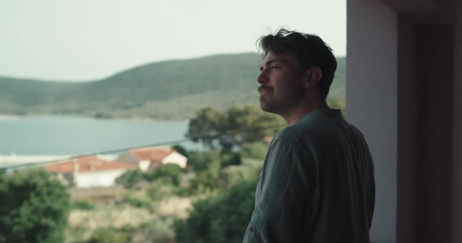 A man walks out on to his balcony and looks out at the shore and hills and nods his head