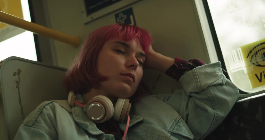 A girl with wine colored hair and headphones around her neck is sitting on a train leaning her head on her hand