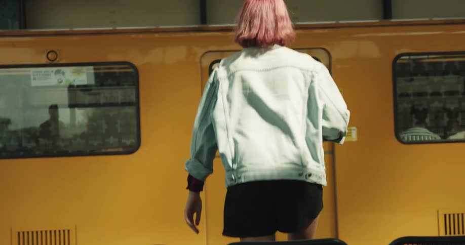 Woman with pink hair denim jacket and black shorts sitting on bench and waiting for train gets on yellow train