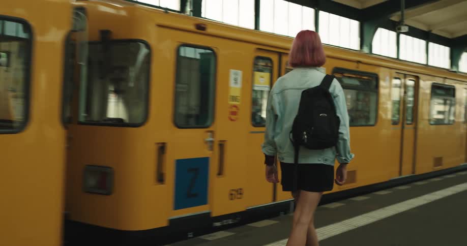 A girl with wine colored hair and wearing a backpack walks up stairs to get on a train