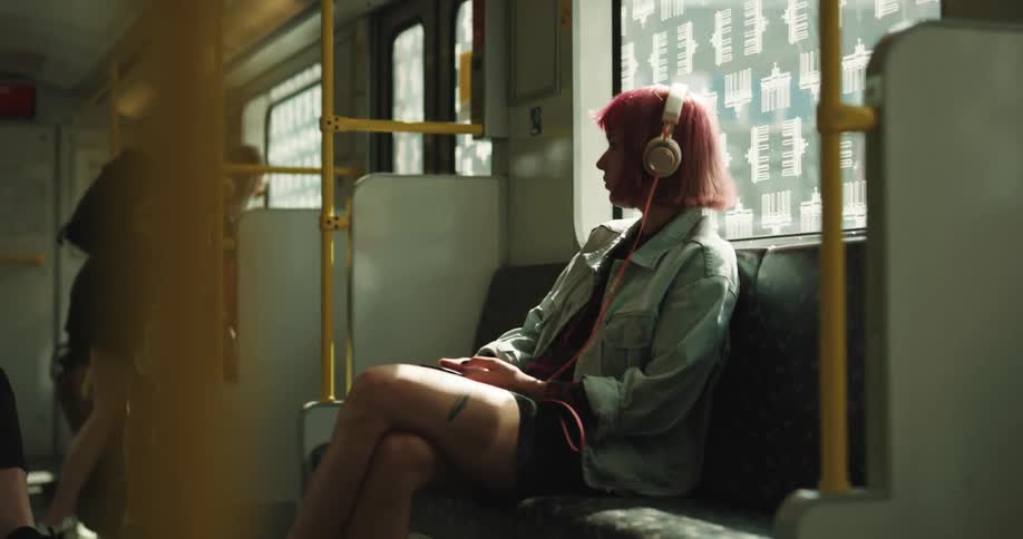 A woman sitting on a train wearing headphones leans forward as another gets up to exit