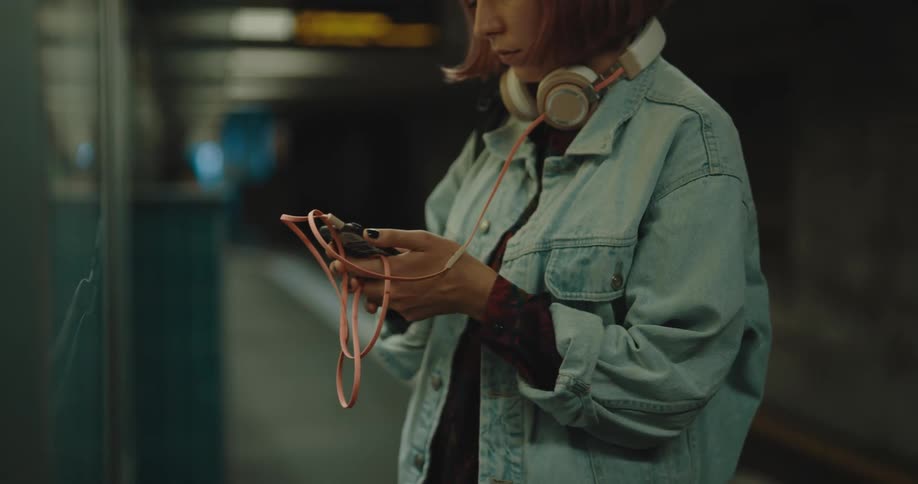 A woman with headphones and a smart phone on a train station with signs