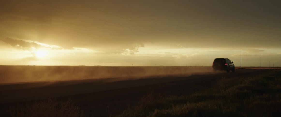 A car is traveling on a desolate dirt road at sunset