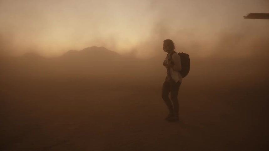 Woman wearing mask and carrying backpack walks through dust storm past short pole with triangular flag