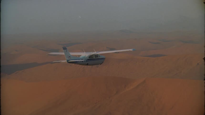 A small plane flies over a barren mountain range on a foggy day