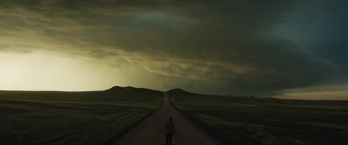 A man is standing on an open road looking at the storm in the sky