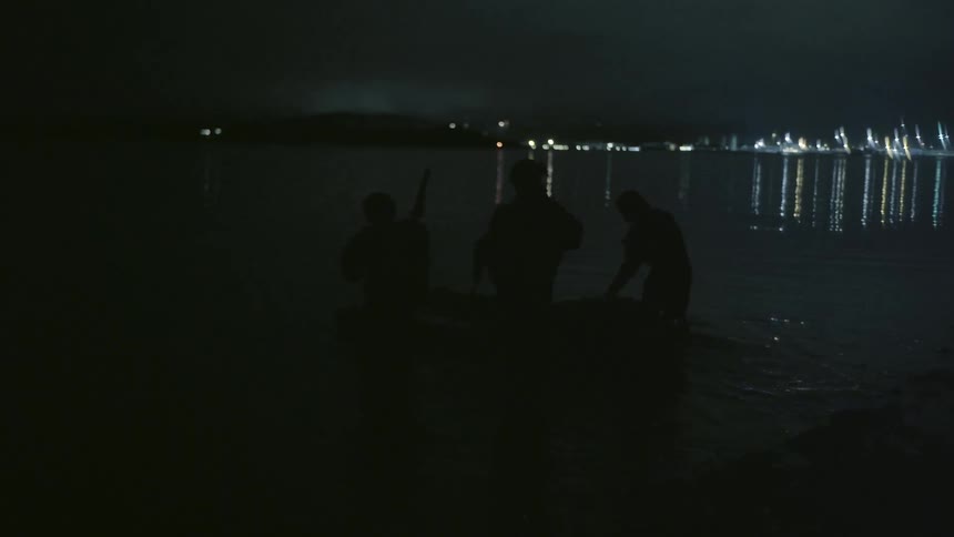 Soldiers guide small boat from shore silhouetted by lights from distant town