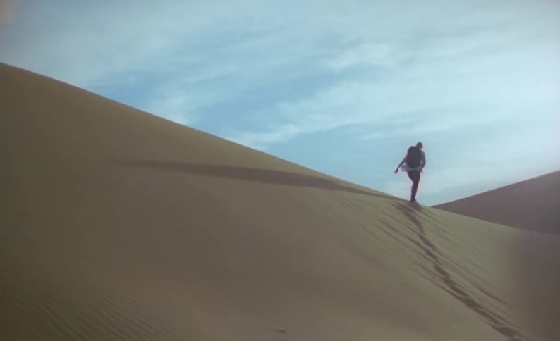 A hiker leaves a trail of foot prints on a sand dune as they climb up the slope to the ridge