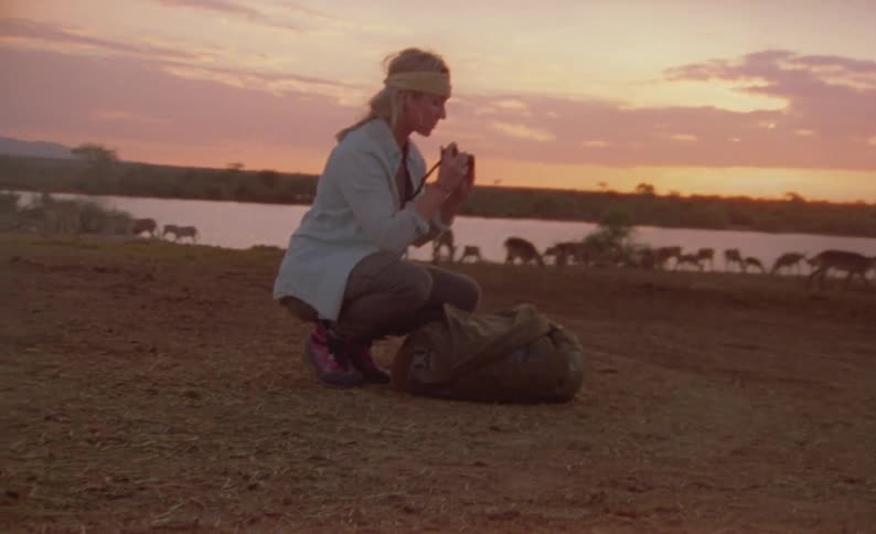 A woman with a backpack and camera taking pictures of animals with the sky and clouds in the background