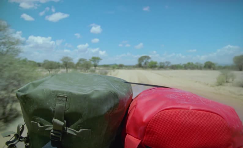 A vehicle with two bags driving in a landscape with dirt and trees with the sky and clouds in the background