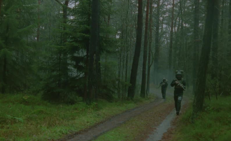 Two men in a paintball battle are walking and patrolling along a dirt road through the forest on a wet and dreary day
