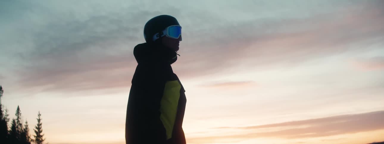 A snowboarder stands on a slope at sunset preparing for a run