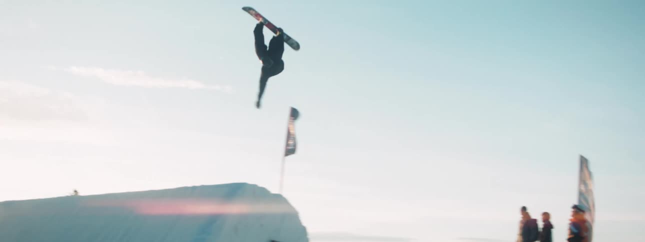 Snowboard rides down slope then up ramp before flipping in midair as people stand and watch