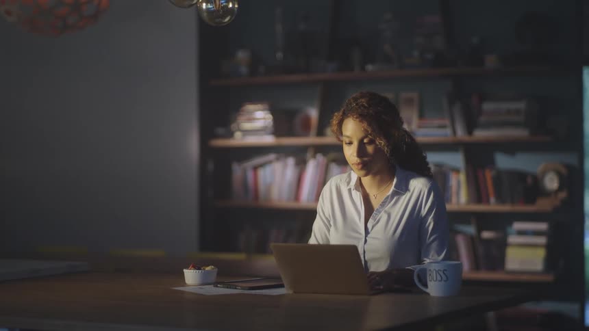 A woman in a room using a tablet with a stylus and typing on a laptop with a bookshelf and a wall in the background