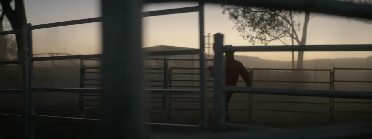 Man in brown jacket jumps over farm fence and closes the fence door at dusk