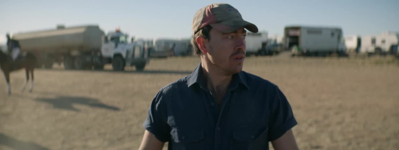 A worried looking man is walking through a dirt lot away from trucks and trailers