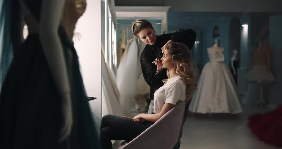 A woman is applying makeup to a prospective bride that is seated in a wedding boutique