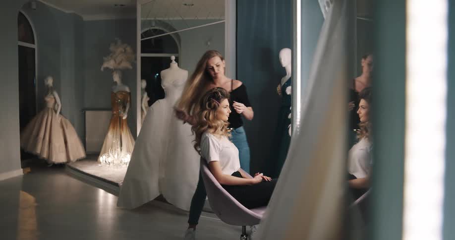 Gowns on mannequins in a design studio where a woman sitting in a chair is having her hair done by a stylist