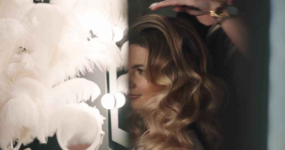 A hair stylist styles the hair of a woman sitting in front of a vanity