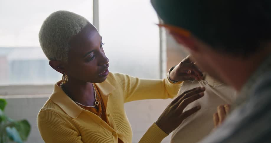 A woman wearing a yellow sweater is talking to a man as she pins fabric to a mannequin