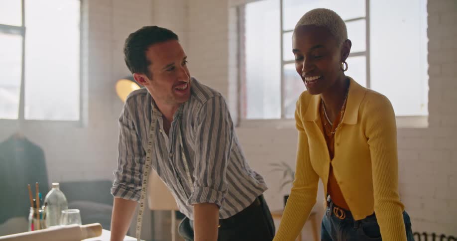 A woman taps on a screen as she and a coworker stand talking at a work table with patterns and designs on it