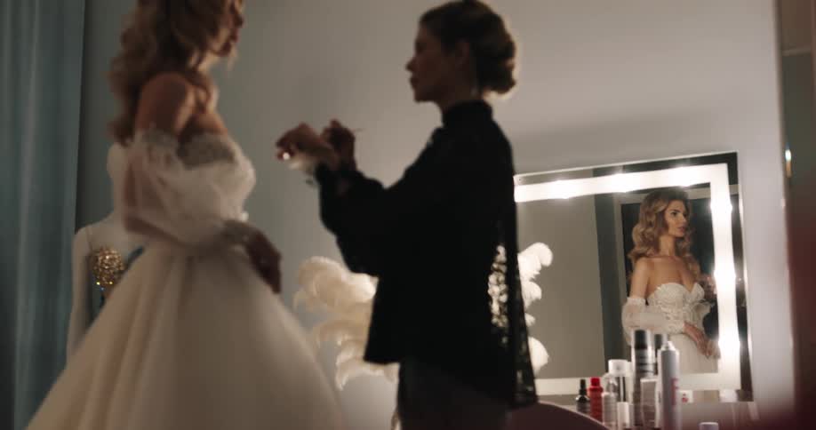 A woman in a room with a wedding dress as another woman applies makeup to her face with a mirror and a wall in the background