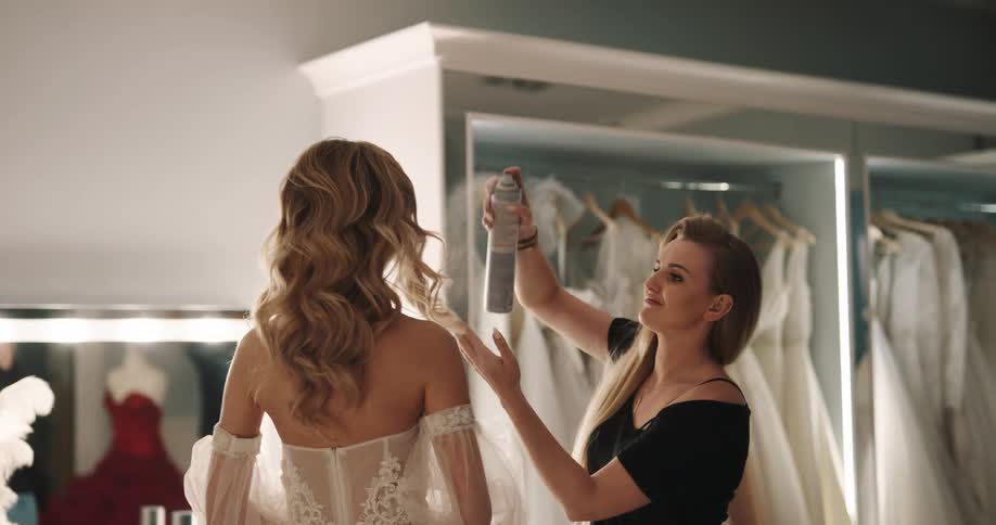 Blonde woman with black shirt sprays womans brown wavy hair wearing wedding dress in bridal shop