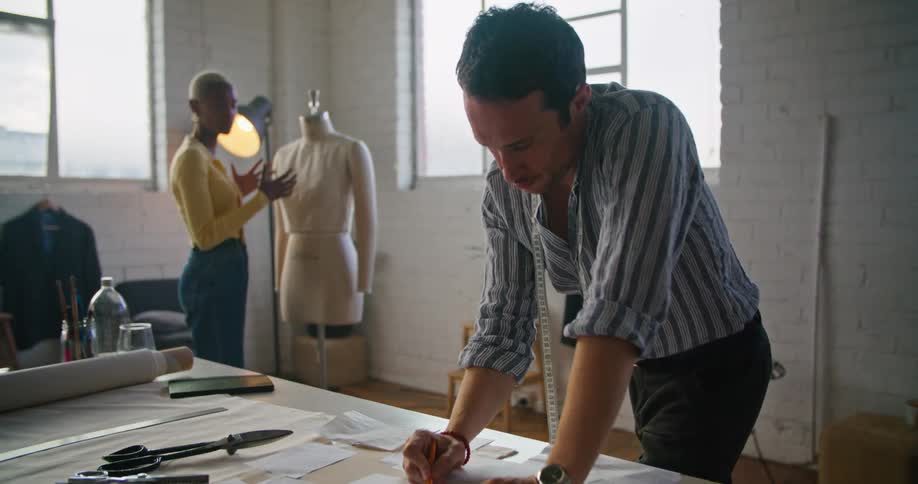 A man sketches at a work table while a woman fits a pattern to a dress form
