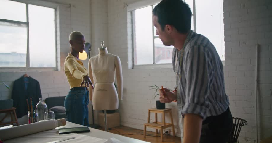 A black woman wearing a yellow sweater and blue jeans is standing near a mannequin explaining something to a man leaning over a desk
