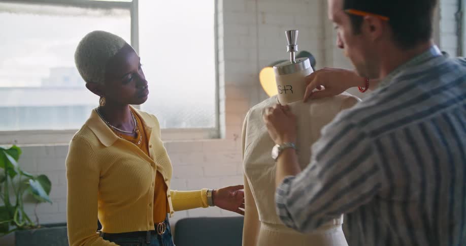 A black woman wearing a yellow sweater is talking to a man in a striped shirt as they adjust clothes on a mannequin