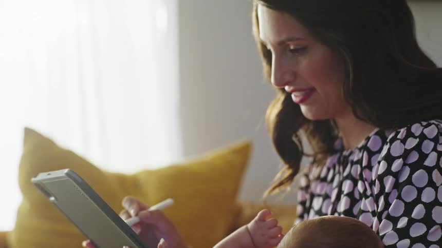 A woman cradles a baby with won arm while using a stylus on a computer tablet as she sits on a sofa in the living room of a house during the day