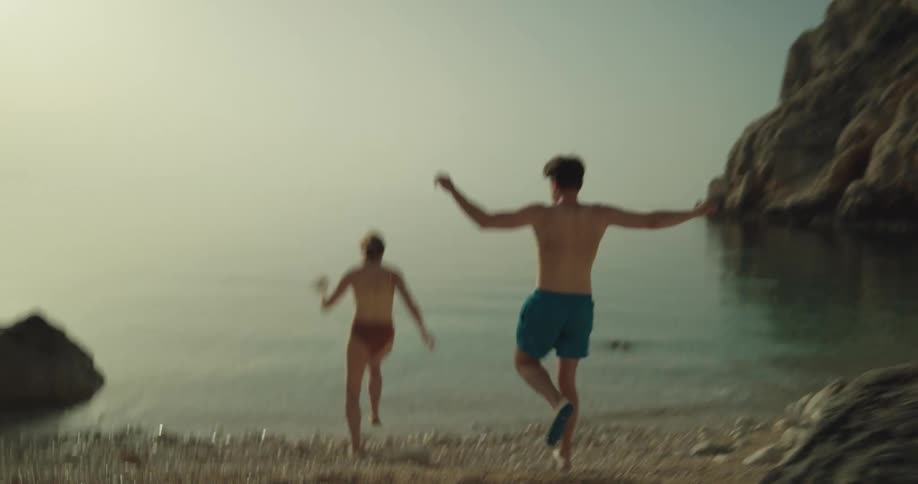 A man and woman on the beach take their shirts off and go running in to the water and laugh and splash one another