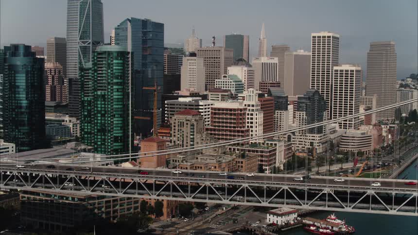 Cars drive across a bridge in front of skyscrapers in a downtown area on the coast
