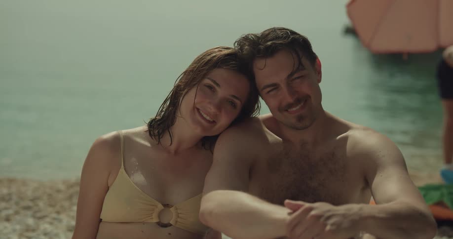 Shirtless man and woman in yellow bikini sits on pebble beach and smile at camera