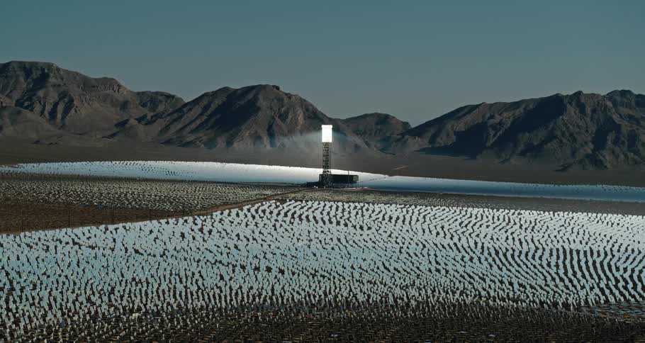 An aerial view of a large array of mirrors with a glowing tower in the middle with a mountain range in the background