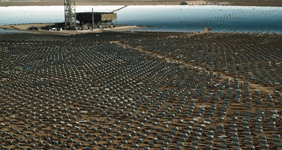 Energy plant with land filled with solar panels and several towers near buildings and mountains in the distance