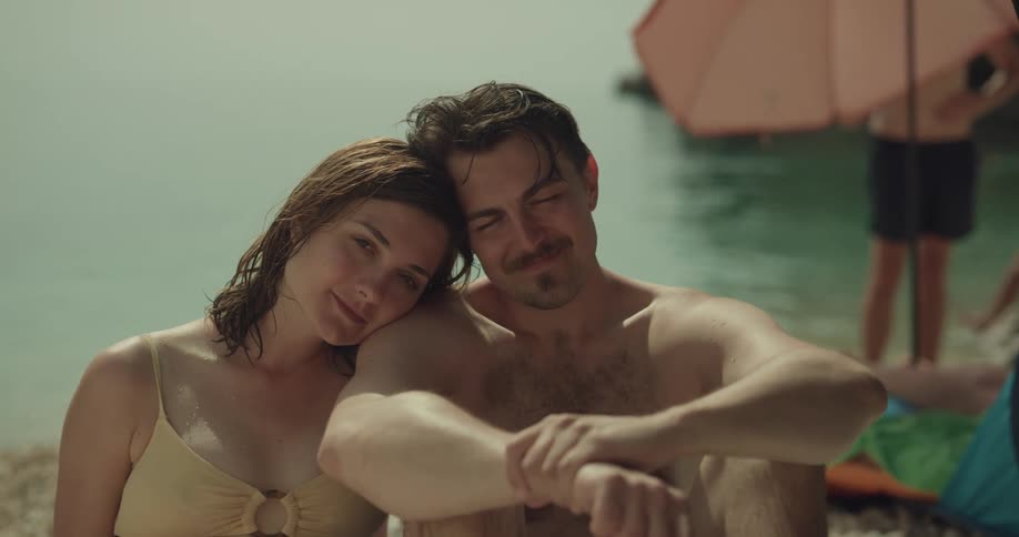 A man and a woman sitting on a sandy beach with people and water in the background