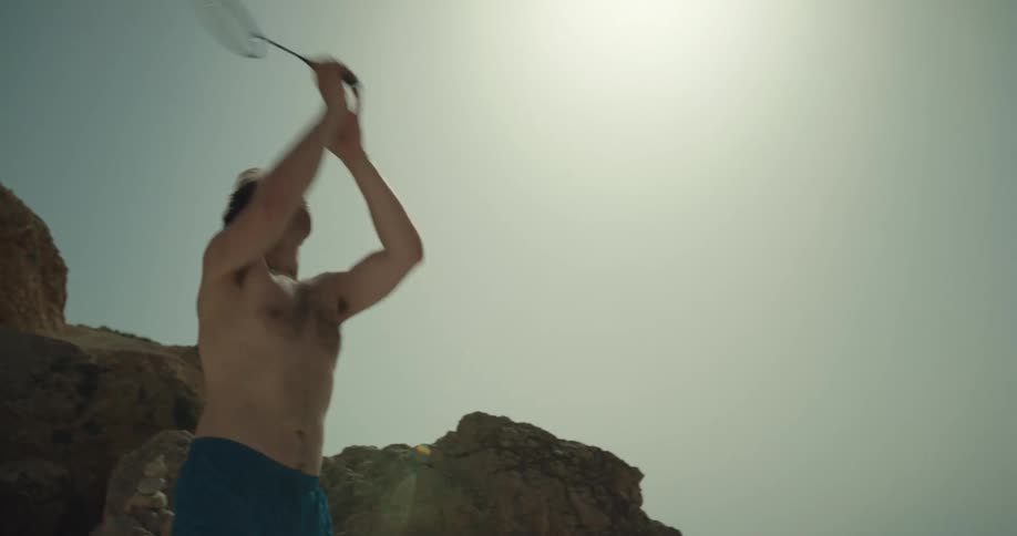 A man is hitting a ball with a badminton racket outdoors by large boulders