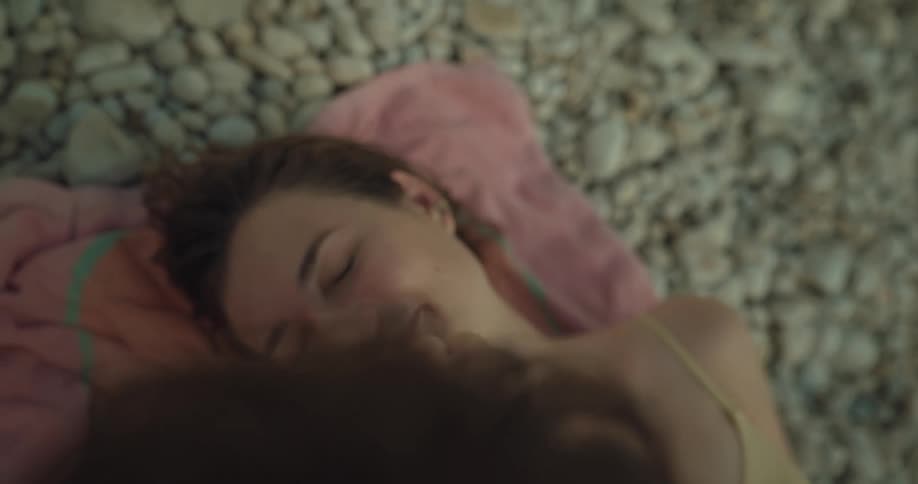 Young woman wearing a swimsuit laying over a towel over pebbles laughing and kissing a man