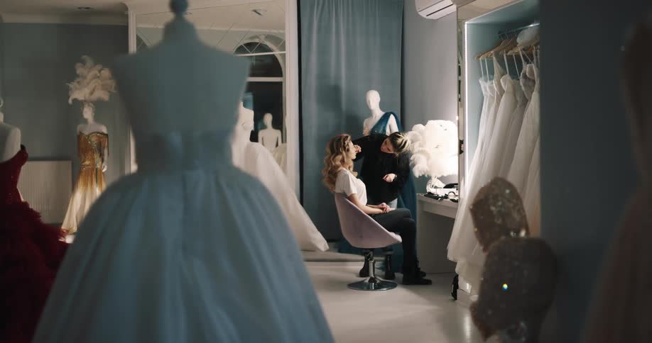 Makeup artist applies makeup to woman sitting in bridal shop displaying wedding dresses
