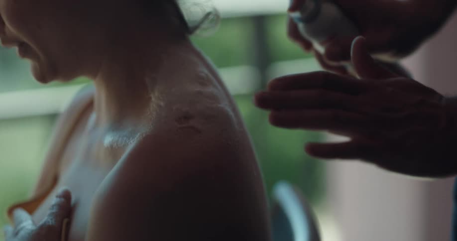 A woman holds onto a towel as a man applies spray on sunscreen to her back