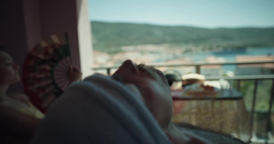 A man leans back and rests in his hotel room of the shore while a woman sits and fans herself