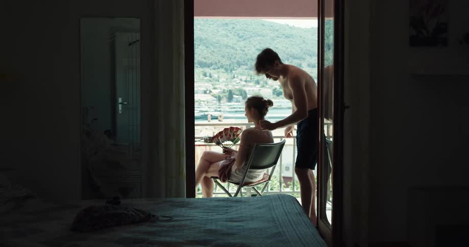 A woman fans herself as her boyfriend applies sunscreen to her back while theyre on the balcony of their hotel room